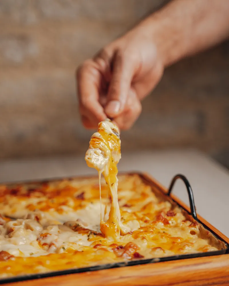 A hand pulls the cheese from a freshly grilled mac and cheese in its dish