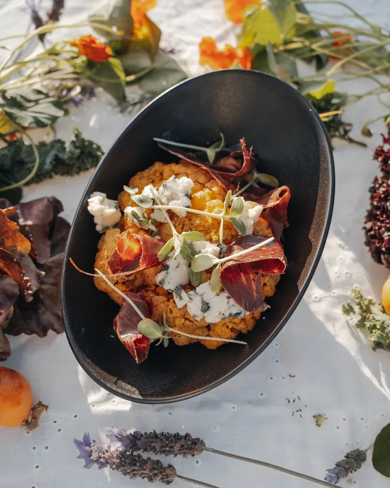 Coliflor asada con queso y jamón en un cuenco negro, rodeada de flores