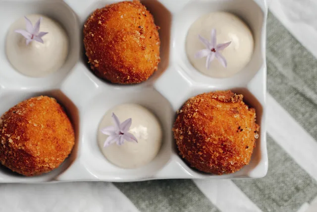 Croquetas en una huevera de cerámica blanca, con salsa blanca y flores lilas
