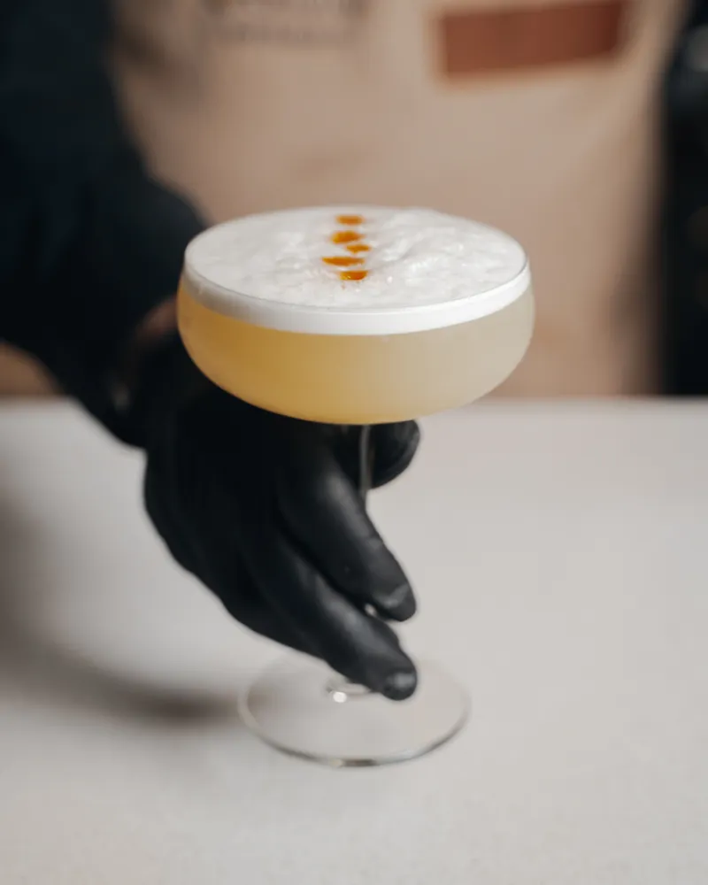Una mano con guante negro sirve un cóctel con espuma blanca y unas gotas naranjas